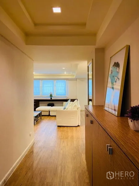 Condominium for rent in Watthana, Bangkok - Hallway with recessed ceiling lights and a wood cabinet leading into a bright living room with large windows.