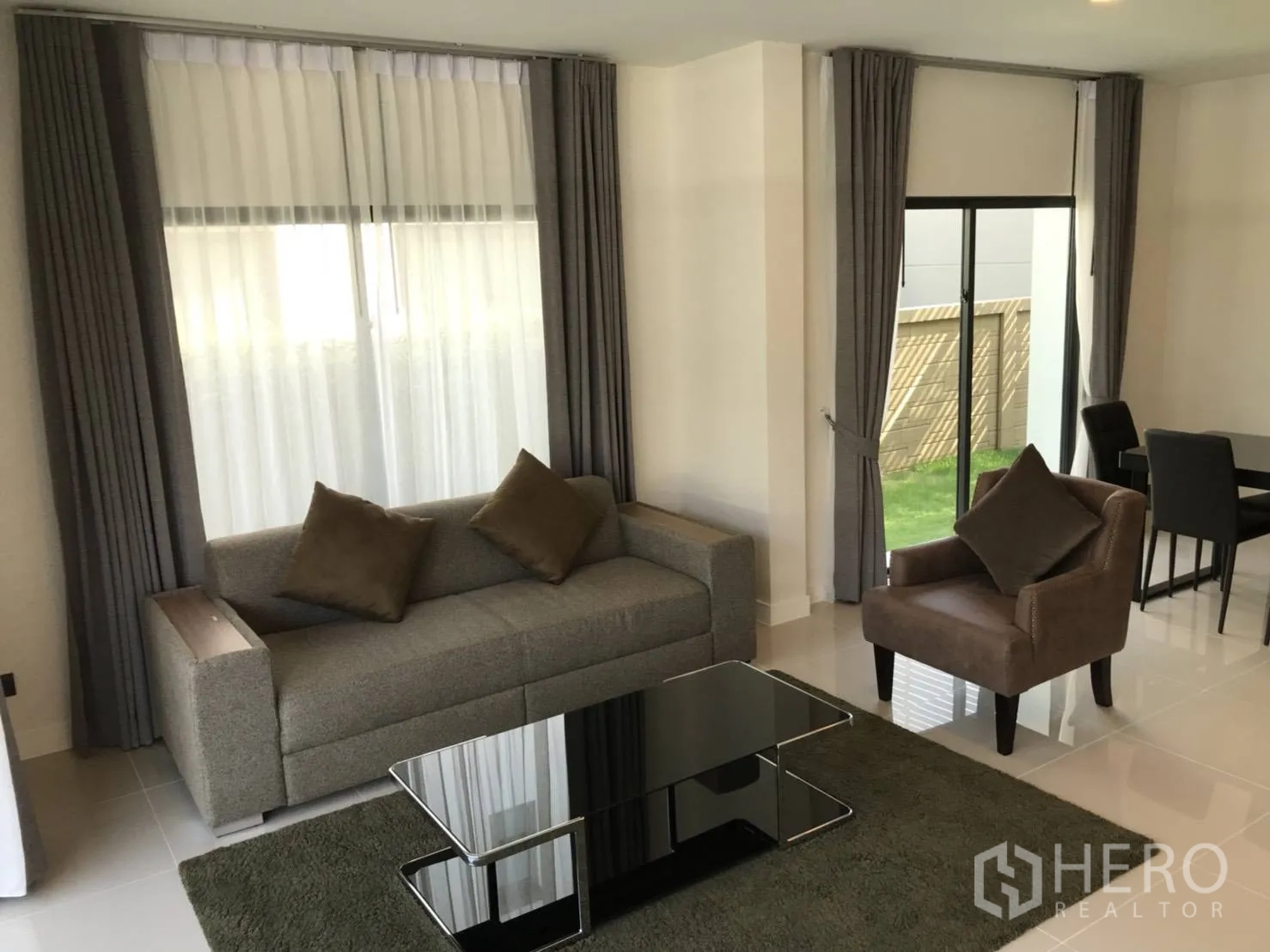 Detached House for rent in Bang Kapi, Bangkok - Bright living room with gray sofa, armchair, glass coffee table and full-height curtained windows.