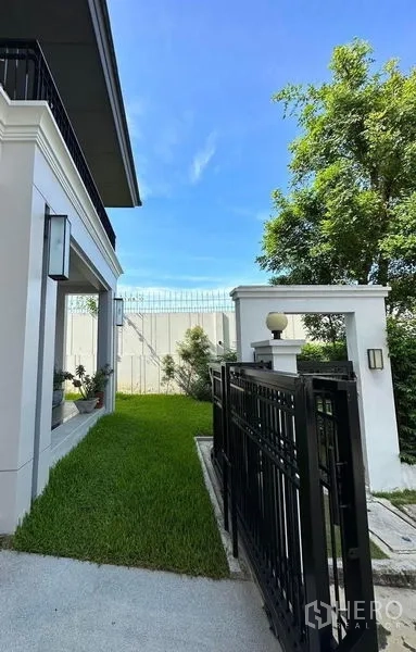 Detached House for sale in Prawet, Bangkok - Side gate and landscaped green yard beside the house under a blue sky.