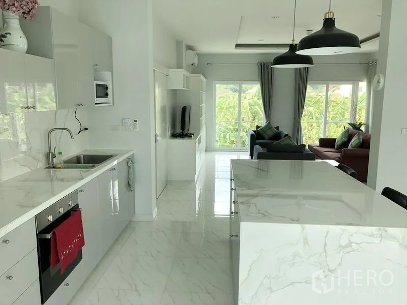 Detached House for rent or sale in Huai Khwang, Bangkok - Long marble-counter kitchen and island opening to a living area with green views from large windows.