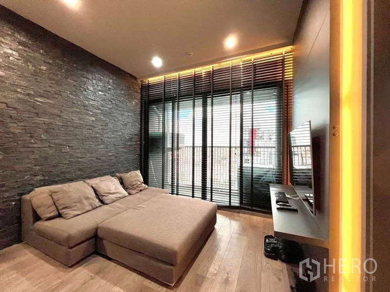 Condominium for rent - Modern living room with a gray sectional sofa, floor-to-ceiling windows with blinds, balcony view, and a dark textured feature wall.