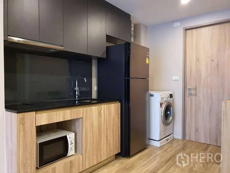 Condominium for rent in Sathon, Bangkok - Compact kitchen with black countertop, upper cabinets, fridge and washing machine.
