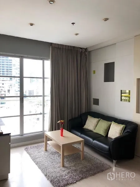 公寓 出租在Khlong Toei, Bangkok - 明亮客厅，落地窗与灰色窗帘，黑色皮沙发与浅木茶几，享城市景观。