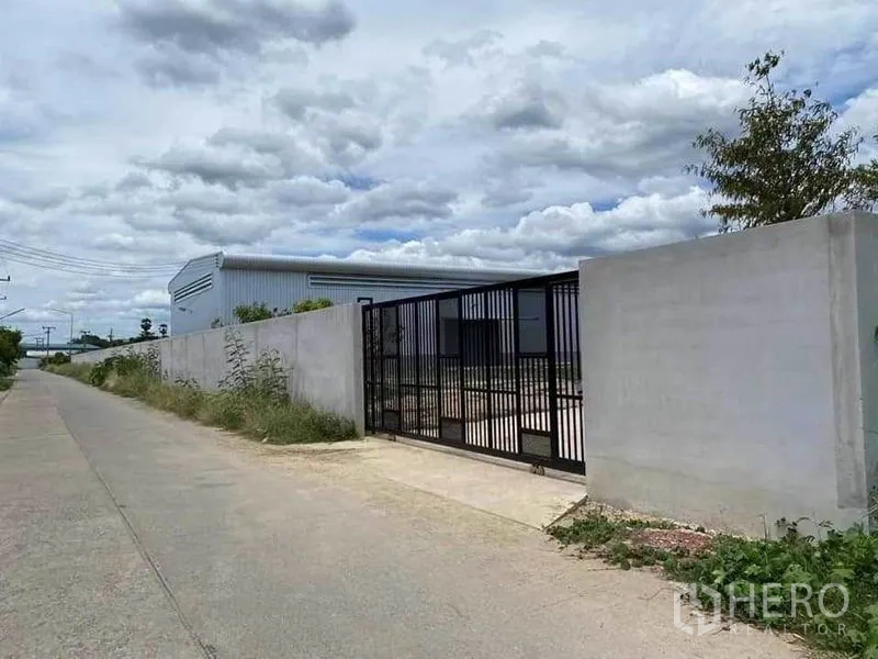 Warehouse for rent in Sam Phran, Nakhon Pathom - Street view of gated warehouse entrance with perimeter wall and access road in Sam Phran.