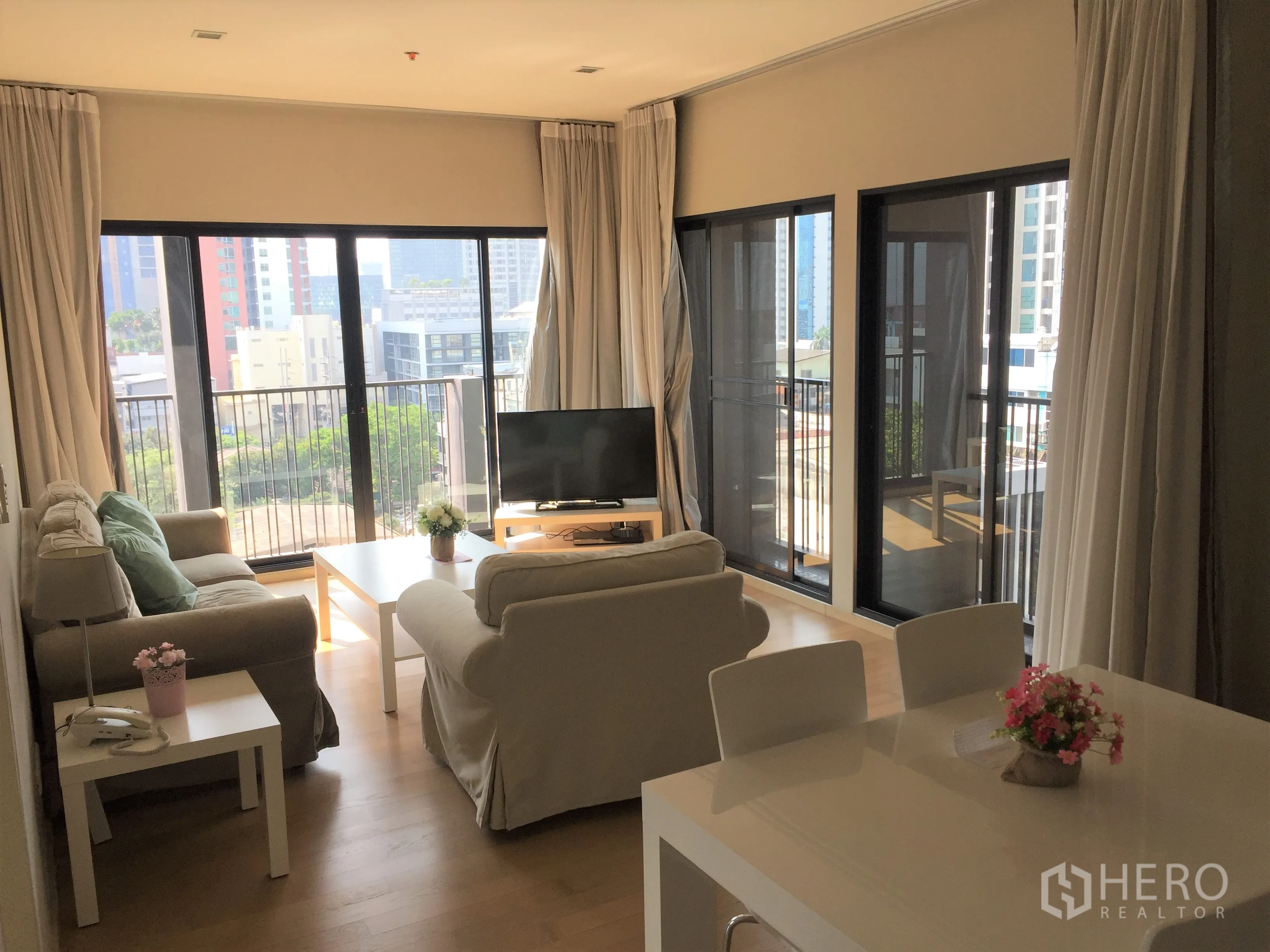 Condominium for rent or sale in Watthana, Bangkok - Sunlit living and dining area with sofas, TV, and multiple balcony doors overlooking Ekkamai.