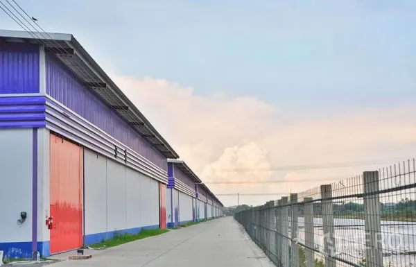 货栈 出租在Pathum Thani - 沿围栏与排水河道的仓库侧视，红门整齐排列，旁有服务道路。