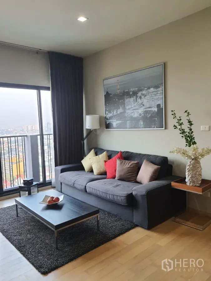 Condominium for rent or sale in Watthana, Bangkok - Living room with gray sofa, colorful cushions, low coffee table and balcony with skyline views.