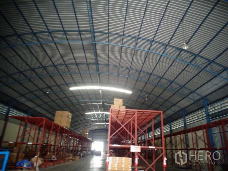 Warehouse for rent or sale in Lat Lum Kaeo, Pathum Thani - Curved corrugated roof with blue trusses and tall red storage racks inside the warehouse.
