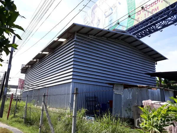 Condominium for rent in Pathum Wan, Bangkok - Metal-clad building with a gabled roof beside a fenced grassy lane and overhead power lines.