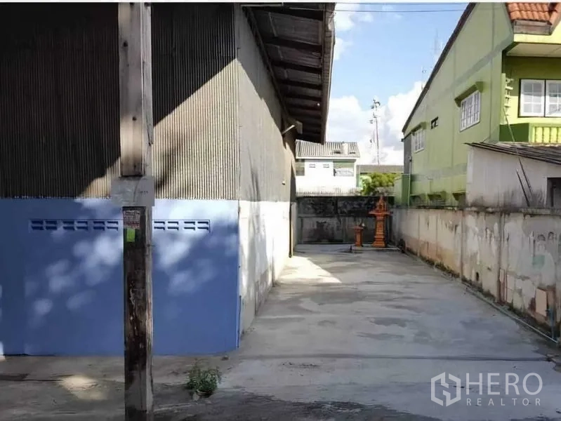 Warehouse for rent in Sai Noi, Nonthaburi - Walled concrete side yard beside corrugated metal warehouse with blue doors