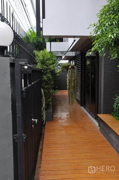 บ้าน ให้เช่าในWatthana, Bangkok - ทางเดินด้านข้างพื้นไม้ ล้อมด้วยต้นไม้และรั้วเหล็กสีดำ