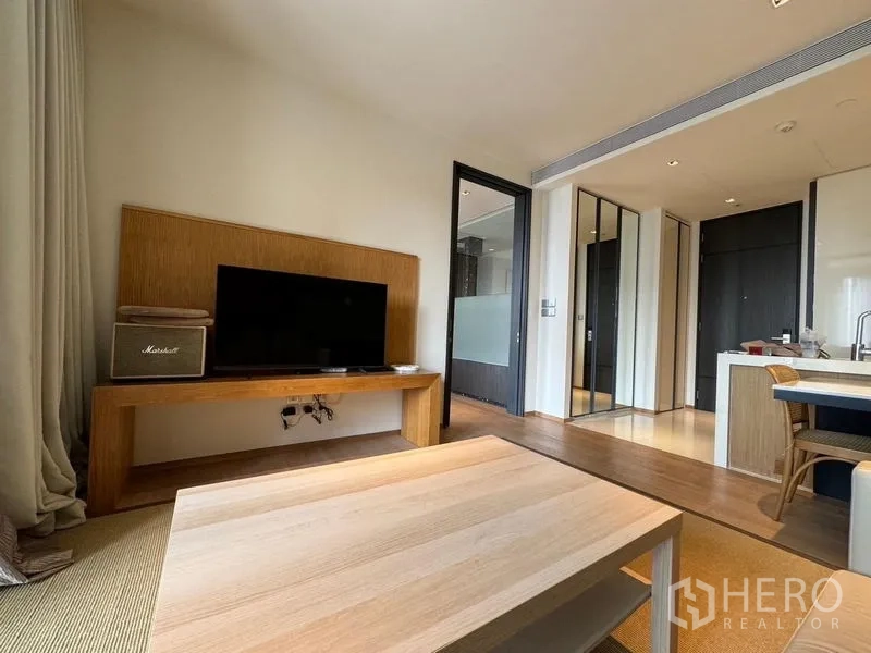 คอนโด ให้เช่า - พื้นที่นั่งเล่นมีชั้นวางทีวี โต๊ะกลาง มองต่อเนื่องไปยังครัวแบบเปิดและโถงทางเข้า