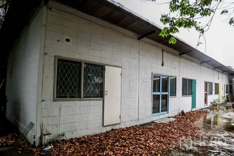Warehouse for rent in Din Daeng, Bangkok - Single-storey auxiliary building with metal roof, several doors and windows beside a leaf-strewn yard.
