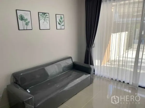 联排别墅 出租在Prawet, Bangkok - 采光充足的客厅，灰色沙发、墙面装饰画与落地窗帘。