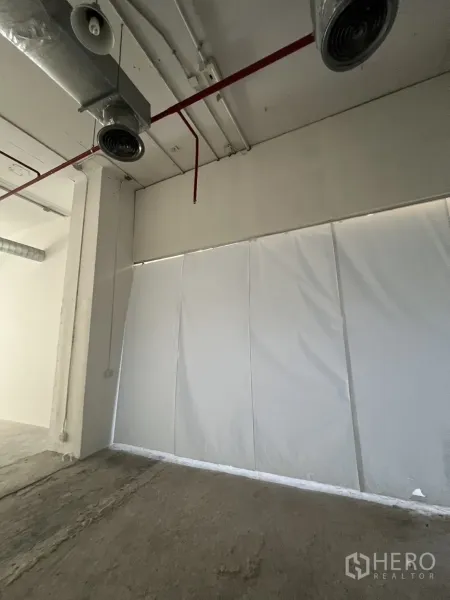 พื้นที่ขายปลีก ให้เช่า - ผนังด้านข้างกั้นชั่วคราว มีท่อสปริงเกลอร์สีแดงและช่องลม พร้อมพื้นคอนกรีตดิบ