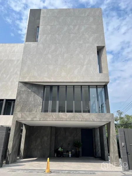 Home Office for sale in Prawet, Bangkok - Modern three-storey home office facade with grey stone cladding and a covered carport under a blue sky.