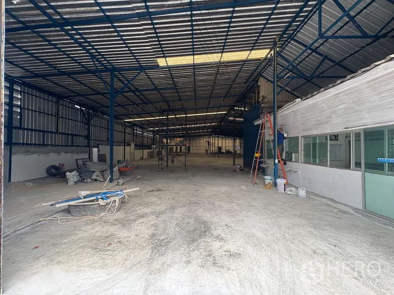 货栈 出租在Prawet, Bangkok - 仓库入口视角，混凝土地面、金属波纹墙，梯具与一侧玻璃隔断办公室可见。