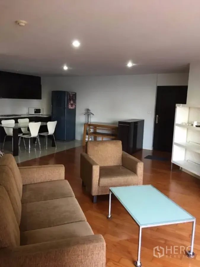 Condominium for rent or sale in Yan Nawa, Bangkok - Open-plan lounge with sofa and armchair facing a kitchenette, shelving unit, and refrigerator.