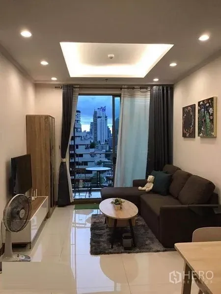 公寓 出租在Watthana, Bangkok - 温馨客厅，沙发、电视与通向阳台的落地门，远眺天际线。