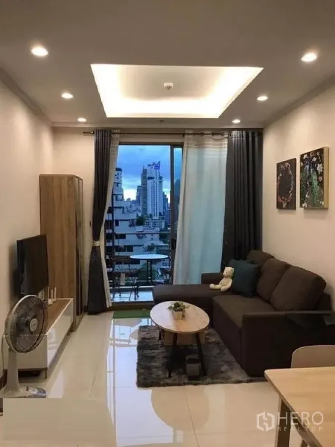 公寓 出租在Watthana, Bangkok - 温馨客厅，沙发、电视与通向阳台的落地门，远眺天际线。