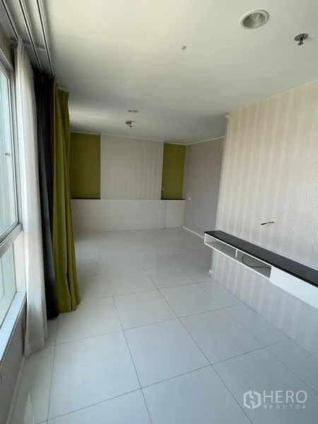 Condominium for sale in Suan Luang, Bangkok - Spacious room with large window, green drapes, wall-mounted media shelf and white tile flooring.