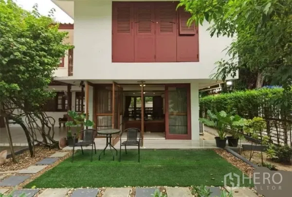 独立式住宅 出租在Watthana, Bangkok - 两层独栋别墅外观，红色百叶窗，落地门通向草坪花园与户外桌椅