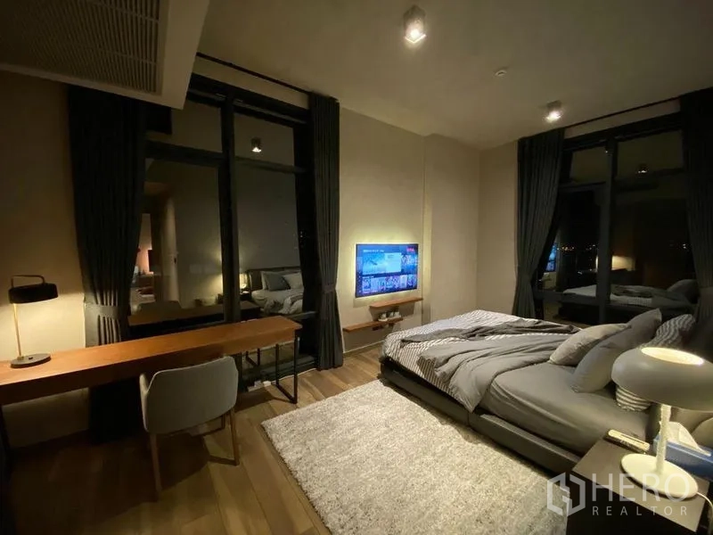 Condominium for rent or sale in Watthana, Bangkok - Spacious master bedroom with large windows, work desk, rug, and wall-mounted television.