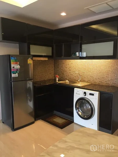 Condominium for rent or sale in Watthana, Bangkok - L-shaped glossy black kitchen with mosaic backsplash, sink, cooktop, fridge, and washing machine.