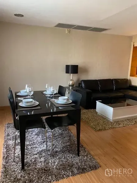 Condominium for rent in Watthana, Bangkok - Open-plan living and dining area with a black dining table, leather sofa, coffee table and rug on warm wood flooring.