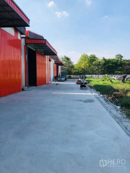 Warehouse for rent in Wang Thonglang, Bangkok - Red and grey metal-clad warehouse with wide concrete loading yard and grassy verge under blue sky.