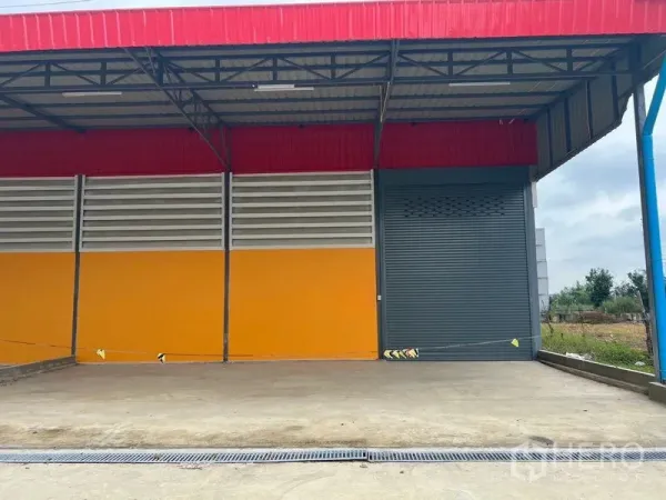 Warehouse for rent or sale in Sai Noi, Nonthaburi - Covered loading dock with a grey roller shutter, drainage channel, and colorful warehouse facade in Sai Noi.