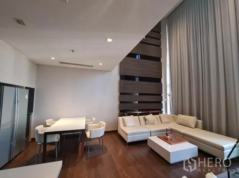 Condominium for rent or sale in Huai Khwang, Bangkok - Open-plan living and dining area with L-shaped sofa, wood floors and tall drapes.