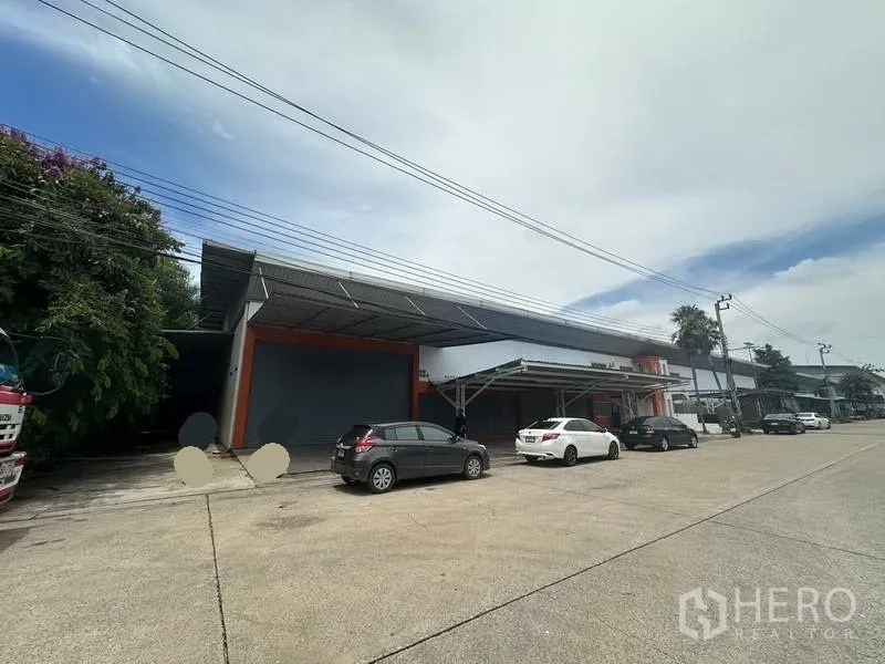工厂 出租在Nonthaburi - 工厂外立面与宽阔路面，设有卷帘门及钢结构雨棚下的停车位。
