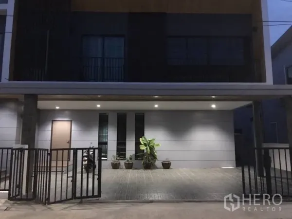 Detached House for rent in Thanyaburi, Pathum Thani - Modern detached house exterior at dusk with covered carport, gated entry, and potted plants in Thanyaburi.