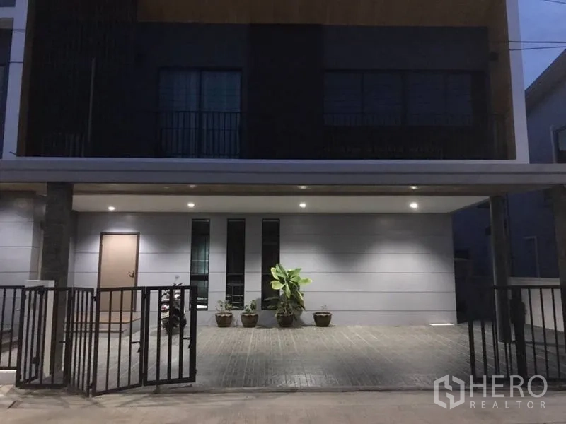 Detached House for rent in Thanyaburi, Pathum Thani - Modern detached house exterior at dusk with covered carport, gated entry, and potted plants in Thanyaburi.