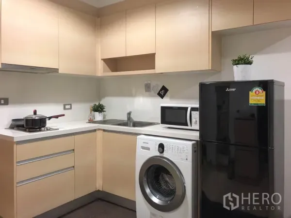 公寓 出租或出售在Watthana, Bangkok - 紧凑L型厨房，带橱柜、水槽、微波炉、洗衣机与黑色冰箱。