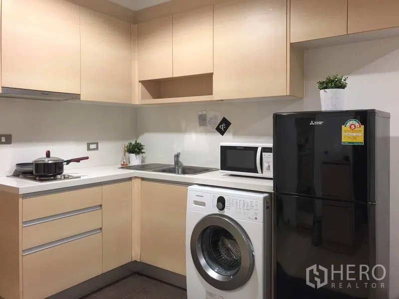 公寓 出租或出售在Watthana, Bangkok - 紧凑L型厨房，带橱柜、水槽、微波炉、洗衣机与黑色冰箱。