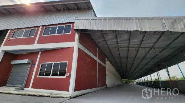 Warehouse for rent in Nonthaburi - Extended metal canopy forming a long covered loading area beside the red warehouse and office block, suitable for truck access.