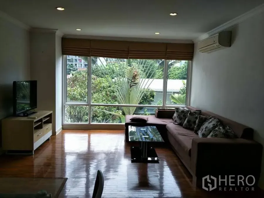 公寓 出租在Khlong Toei, Bangkok - 客厅采光充足，木地板，L形沙发与玻璃茶几，电视柜，大窗户眺望绿景