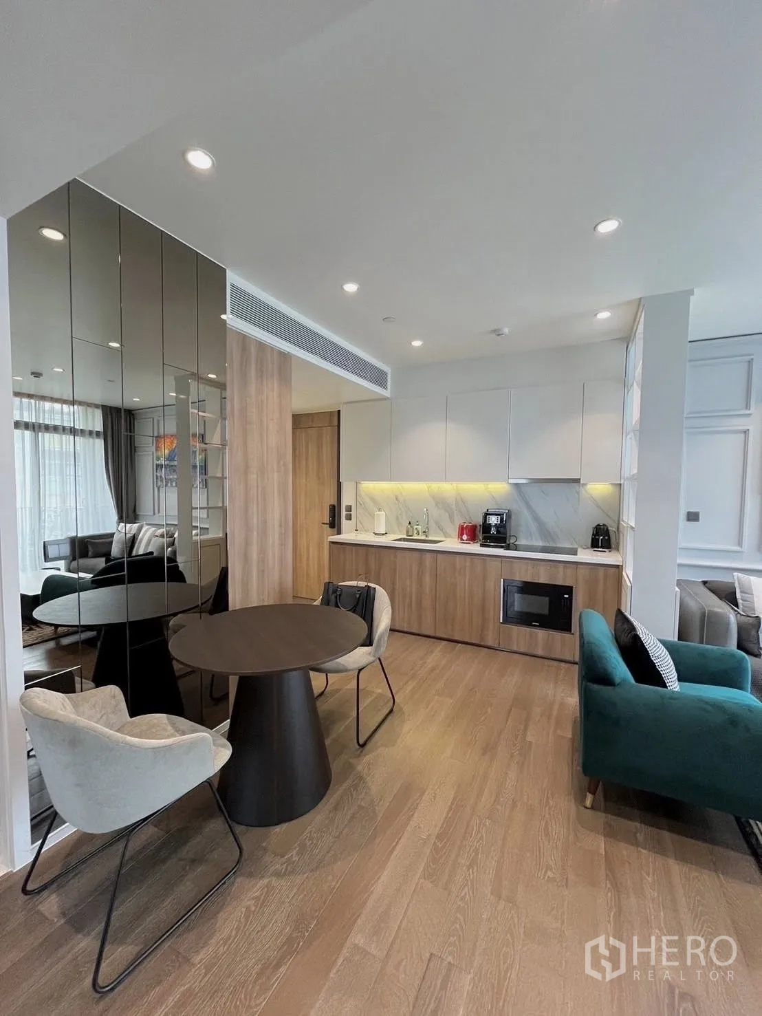 Condominium for rent in Watthana, Bangkok - Open-plan living area with round dining table, mirrored wall panels, wood floors, compact kitchenette with built-in appliances, and a teal sofa.