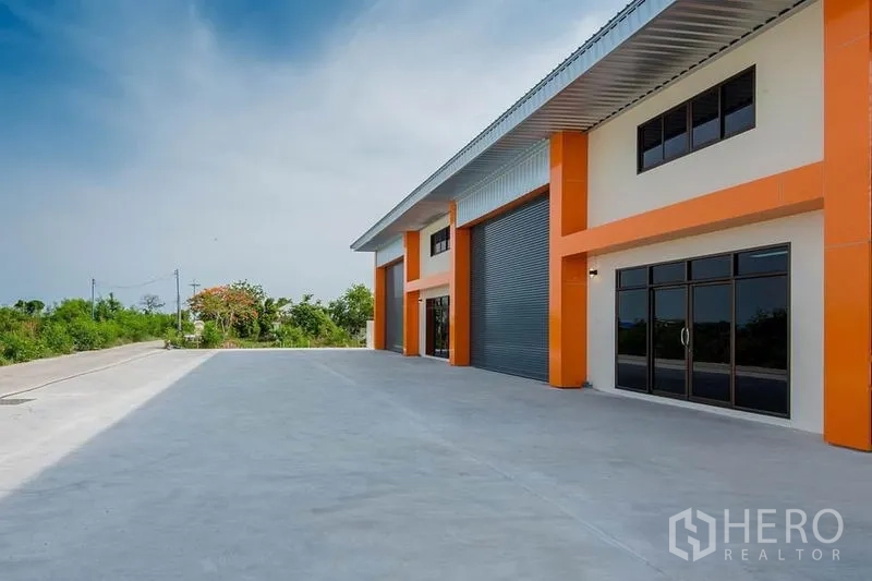 Factory for sale in Bang Sao Thong, Samut Prakan - Side view of the factory showing glass-fronted office entrances, roller shutter bay and expansive driveway with greenery.