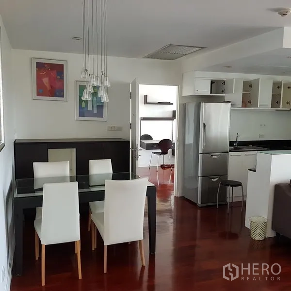 公寓 出租在Pathum Wan, Bangkok - 餐区配玻璃餐桌与白色餐椅，吊灯点缀，毗邻现代厨房与小型书桌区。