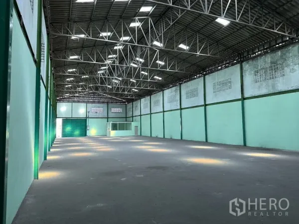 Factory for rent in Bang Bo, Samut Prakan - Spacious factory interior with high steel-truss roof, skylights, concrete floor, and a loading door ahead.