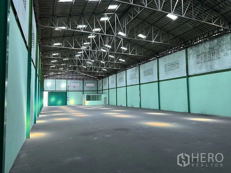 Factory for rent in Bang Bo, Samut Prakan - Spacious factory interior with high steel-truss roof, skylights, concrete floor, and a loading door ahead.