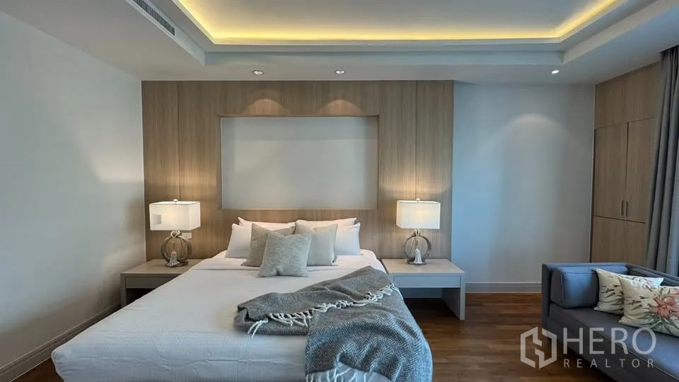 คอนโด for rent in Pathum Wan, Bangkok - ห้องนอนหลักเตียงขนาดใหญ่ ผนังลายไม้ โต๊ะข้างเตียงพร้อมโคมไฟ ตู้บิลต์อิน และเพดานซ่อนไฟพร้อมโซฟา