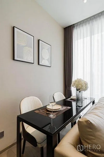 Condominium for rent in Watthana, Bangkok - Compact dining nook for two with a black glass table beside tall sheer curtains.