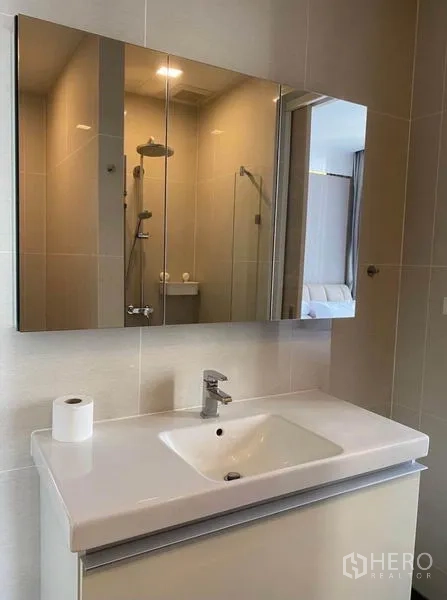 Condominium for rent in Khlong Toei, Bangkok - Modern bathroom vanity with large mirrored cabinet and a glimpse of the glass shower.