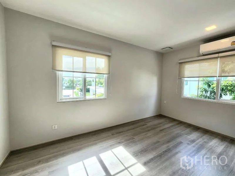 Detached House for rent or sale in Bang Phli, Samut Prakan - Sunlit empty bedroom with wood flooring, corner windows and an air conditioner.