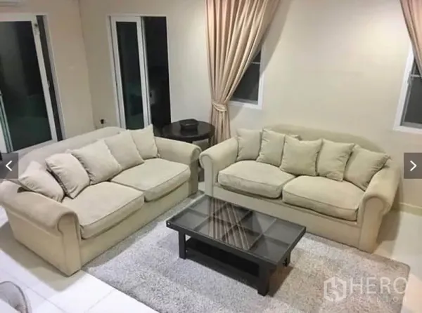 Townhouse for rent or sale in Suan Luang, Bangkok - Bright living room with two beige sofas, glass coffee table, curtains, and sliding doors.