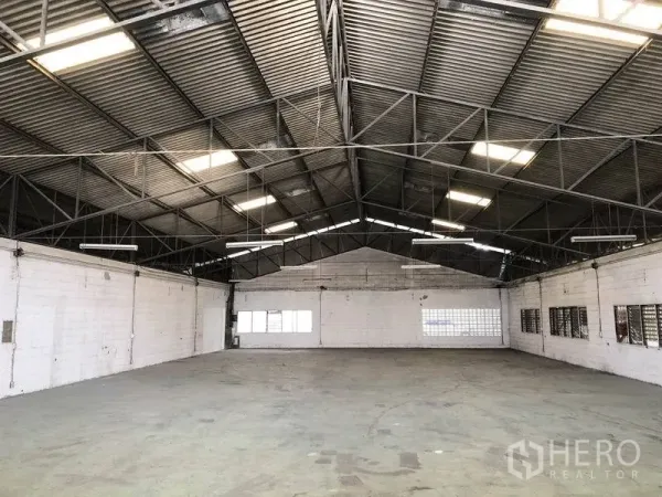 Warehouse for rent in Mueang Pathum Thani, Pathum Thani - Wide‑angle view of an open‑plan warehouse hall featuring a high gable roof, hanging lights and multiple windows for airflow.